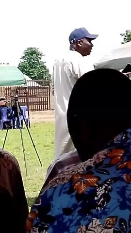 The video shows an outdoor event with several people. In the foreground, the back of a person's head is visible, wearing a blue and orange patterned shirt. Further back, a man in a white robe and a blue baseball cap stands facing away from the camera. To the left of him, another man in a blue shirt and a blue baseball cap is seen holding a device and looking towards a tripod with a camera attached. There are green tents and a wooden fence in the background, along with some trees. The ground appears to be covered in grass. The lighting suggests it is daytime.