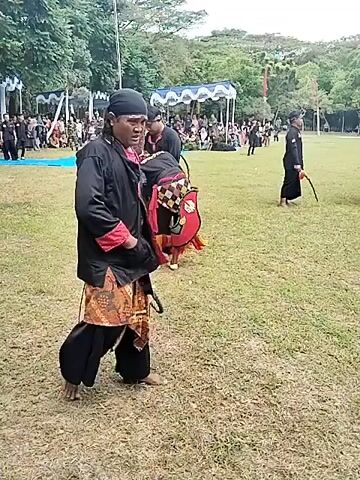 The video captures an outdoor traditional performance, likely a Kuda Lumping (horse dance) event, taking place in a large grassy field in Puro, Indonesia. The sky is overcast, suggesting daytime.

Several individuals are involved in the performance. At the beginning, a man with long hair, wearing a black shirt, red undershirt, brown patterned skirt-like garment, and black pants, walks barefoot while holding a coiled rope. He appears to be a shaman or handler.

Subsequently, a performer dressed in a traditional horse costume (Kuda Lumping), primarily red and black with decorative elements, engages in a dance. Another man in black attire and a head covering guides or interacts with the "horse" performer. Other individuals in similar black traditional clothing are also present in the field, some observing and others participating.

In the background, there's an audience gathered under white tents. A tall pole with what appears to be the Indonesian flag is visible. Lush green trees and distant hills complete the outdoor setting. The overall atmosphere suggests a cultural celebration or demonstration. No discernible text is visible on signs or banners.