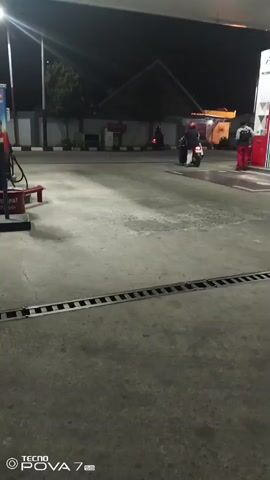 At night, a person on a scooter drives through a gas station. Another person stands by a pump. The ground is concrete and wet in places. There are gas pumps and a building in the background.