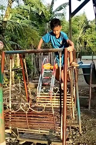 A young boy wearing a blue shirt and shorts stands on top of a rusty metal swing set in an outdoor area with green vegetation. The swing set is multi-colored with red, green, yellow and teal paint. Behind the swing set, a rusty slide and other playground equipment are visible. The boy is balanced on the top bar of the swing set, looking down. The ground appears to be a mixture of dirt and small gravel. The background consists of trees and plants, suggesting a tropical or semi-tropical environment. The video is taken in daylight.