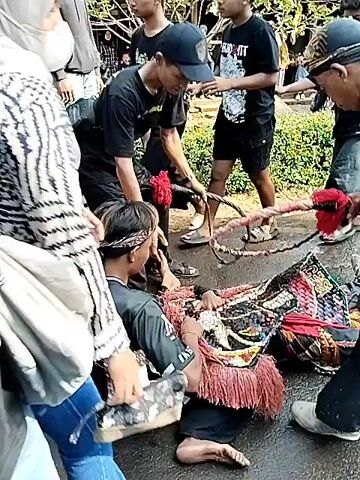 A group of people are gathered around what appears to be a performance or ritual. In the foreground, a person in a black shirt with "BLEDUX IRENG" written in yellow on the back is visible from behind. In the center of the image, several individuals in black shirts and headbands are kneeling and handling a person dressed in a costume with red and pink fringe, possibly a mask or headpiece. Another person in a black shirt with "LENTRENG" written on it is also present. In the background, more people are standing and observing, with trees and a rural landscape visible. The ground is wet, suggesting it may have rained or is a muddy area. Some individuals are wearing masks. The overall scene suggests a cultural event or performance in an outdoor setting.