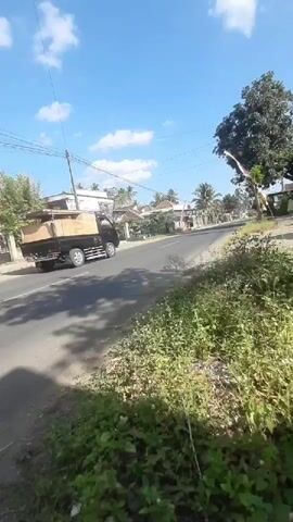 A black truck is seen driving down a paved road in a rural area. The truck is carrying a load on its flatbed. The road is lined with vegetation and houses. The sky is blue with scattered clouds. The video appears to be shot from the side of the road, looking towards the truck as it drives away.