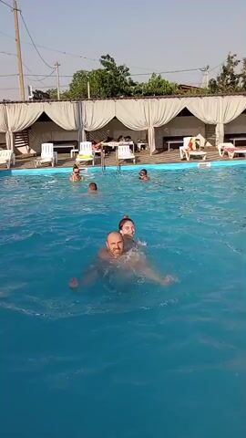 This is a short, horizontal video of a man and a woman in a swimming pool. The man is in the foreground, holding the woman on his shoulders. He is bald and smiling, and the woman has her arms around his neck and is also smiling. They are both splashing water. In the background, there are several other people swimming in the pool. There are also lounge chairs and cabanas along the side of the pool, suggesting it is a public pool or resort. The sun is shining, indicating it is daytime, and the sky is clear. The overall mood is happy and relaxed. There is no visible text in the video.