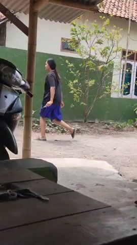 A person wearing a blue skirt and a dark top walks away from the camera. They are walking in front of a green wall with a tree and a window. A motorcycle is parked on the left side of the frame. A wooden table is in the foreground.