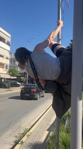 A person is performing a pole dance on a utility pole next to a street. The person is wearing a grey tank top, black shorts with white stripes, and sneakers. They are holding onto the pole with their hands and their legs are extended in various positions. Cars are driving past on the street, and there are buildings and trees in the background. The sky is blue and clear. The video appears to have been filmed during the day.