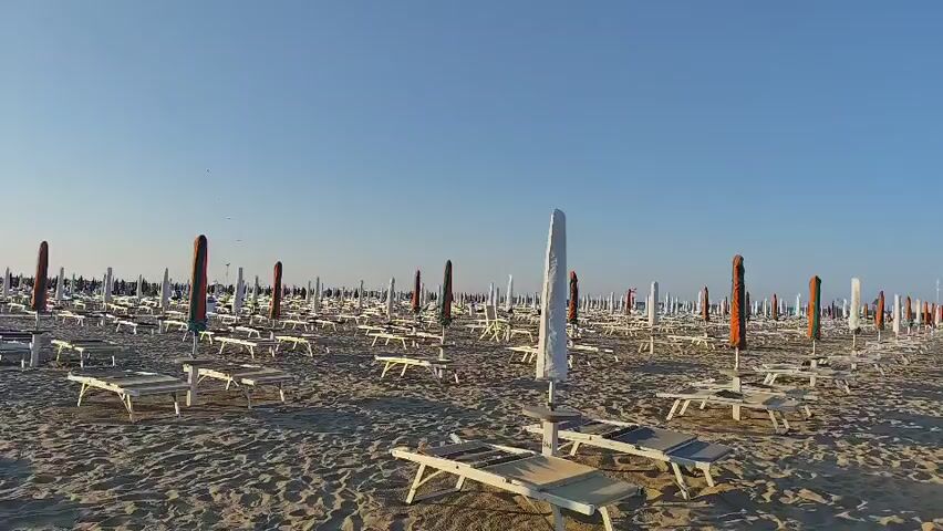 A wide shot shows a vast expanse of a sandy beach with rows of empty lounge chairs and closed umbrellas under a clear blue sky. A white boardwalk runs diagonally across the sand from right to left. Several flags are planted along the boardwalk, and a trash receptacle is visible near its start. The sun casts long shadows across the beach. The scene is peaceful and devoid of people.