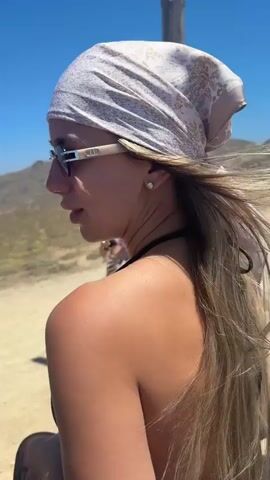 The video opens with a close-up of a woman wearing sunglasses and a headscarf, her back to the camera. The scene then shifts to show a second woman, also wearing sunglasses and a headscarf, standing in the background and gesturing with her hands. The camera moves inside a convertible car, focusing on the hands of the passenger. The video then shows the two women standing outside the car, with a lighthouse and another convertible in the background. The women are smiling and appear to be enjoying themselves. The environment is sunny and appears to be a coastal area with dry, sandy terrain.