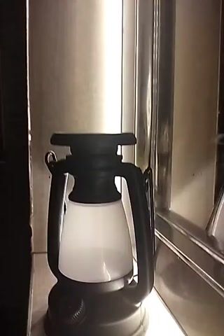 A close-up, still shot of a black and white camping lantern is presented. The lantern, with a matte black frame and a frosted white glass shade, is lit from within, casting a soft glow. The background is blurred, with hints of metallic surfaces and a vertical bright light source. A dial is visible at the base of the lantern.