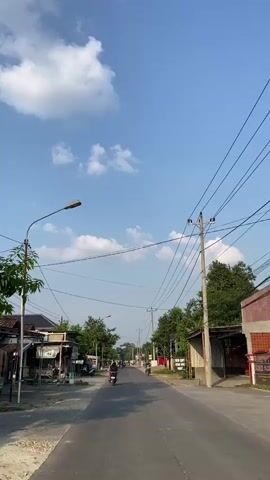 This is a vertical video of a road in a semi-urban or rural setting. The sky is blue with scattered white clouds. A road runs through the center of the frame, with buildings and trees lining both sides. There are power lines and utility poles visible throughout the scene. Several motorcycles are seen on the road, with riders on them. Some of the buildings appear to be shops or small businesses. The lighting suggests it is daytime, likely late morning or afternoon.