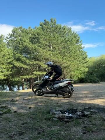 The video shows two parked motorcycles in an outdoor setting with a wooded area and a lake in the background. One motorcycle is a white scooter, and the other is a dirt bike with "AVANTIS" written on it. A person wearing a helmet and protective gear is sitting on the dirt bike, facing away from the camera. To the left of the motorcycles, a person wearing a backpack is walking away from the camera. In the foreground, there is a fire pit with debris. The scene is illuminated by natural daylight.
