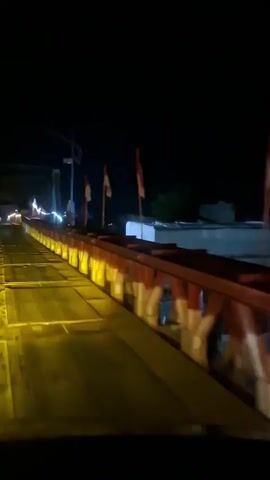 The video shows a dark night scene looking forward from inside a vehicle crossing a bridge. The bridge is illuminated by yellow lights along its sides, and there are red and white flags on poles along the railing. The road surface appears to be made of metal plates. In the distance, there are other lights and some buildings visible. The vehicle is moving forward across the bridge.