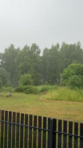 The video shows a heavy rain falling on a green landscape with trees in the background. A wooden fence runs along the bottom of the frame. The sky is overcast. The rain is clearly visible as streaks against the green trees. There are no people or animals visible in the video. The scene appears to be a rural or semi-rural area. The fence is dark brown and appears to be made of wood. The grass in the foreground is green and appears to be somewhat wet from the rain.