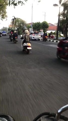 The video shows a street scene from the perspective of someone riding a motorcycle. The camera is pointed forward, capturing the road ahead and the traffic. Several other motorcycles and a few cars are visible. Most of the people in the video are wearing helmets. The road appears to be paved, with a white line marking the lane. The surroundings include trees and buildings, suggesting an urban or suburban environment. The lighting indicates it is daytime, possibly late afternoon or early morning, with some overcast sky. The motion of the camera suggests continuous movement, with a slight blur due to speed.