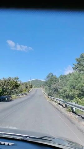 The video shows a view from inside a vehicle, looking out through the windshield. The vehicle is traveling on a paved road, which appears to be a two-lane highway with a guardrail on the right side. The road is surrounded by lush green trees and foliage on both sides. In the distance, a large hill or mountain is visible under a clear blue sky with a few white clouds. The scene suggests a sunny day and a journey through a natural, possibly rural or mountainous area.