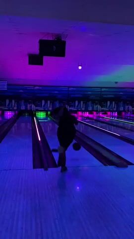 A woman in a black dress and orange shoes walks down a bowling lane. The bowling alley has neon lights that are blue and pink. The ceiling is purple and there are speakers mounted on it. The woman is looking towards the right side of the frame.