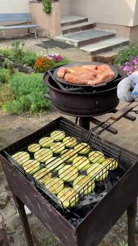 This is a close-up video of a barbecue in a backyard setting in Rezekne, Latvia. The scene focuses on two grills. The top grill, a large flat circular pan, is cooking several pieces of seasoned salmon. The bottom grill contains a metal basket filled with sliced zucchini, which is also being grilled. The salmon appears to be just starting to cook, with some sizzling sounds audible.

The background shows a house with a concrete porch and steps leading up to a door. A light-colored bench is visible in the yard, as well as flower beds with colorful blooms, suggesting it's springtime or summer. Lush green foliage also surrounds the area, hinting at pleasant weather. The lighting suggests it's daytime, likely late afternoon or early evening.

A dog can be seen walking in the background near the stairs, adding a touch of domesticity to the scene. The overall atmosphere is one of a relaxed outdoor meal preparation, evoking a sense of leisure and enjoyment. No text is visible in the video.