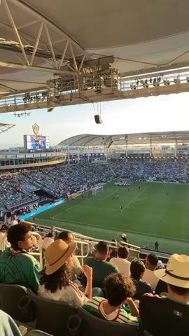 A high-angle medium shot shows a soccer stadium filled with spectators. The sun is setting, casting a warm glow over the scene. The field is green and appears to be in good condition. Numerous players in white uniforms are gathered in a line in the center of the field. Spectators are seated in the stands, some wearing hats and others cheering. The stadium has a large roof structure and various light fixtures. The overall atmosphere appears to be one of excitement and anticipation.
