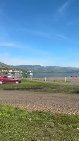A red car is parked on the side of a dirt road near a grassy area. In the background, there is a body of water, possibly a lake or river, with mountains in the distance. The sky is clear and blue. There are a few people walking around on the shore of the water. The overall scene appears to be a scenic outdoor location on a sunny day.