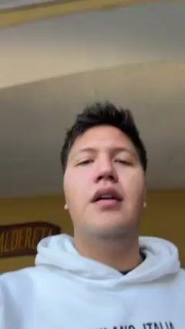The video shows a medium shot of a young man with short, dark hair and light skin. He is wearing a white hoodie with text on it. The camera is held at a slightly upward angle, and the man's head is centered in the frame. He looks around, mostly upwards. In the background, there is a partially visible yellow wall with a sign and a section of a building overhang. The video quality is decent, with natural lighting. The man's expression is neutral to slightly contemplative. The camera appears to be handheld, leading to slight movements.