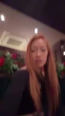 A woman with long red hair sits facing the camera in what appears to be a restaurant. She wears a black shirt with a cutout at the chest. Behind her are red roses and a dark wood paneled wall. An air conditioning unit is visible on the ceiling. She pouts her lips, smiles, then holds up her hands, showing her bracelets, and runs her hands through her hair. Music plays in the background.