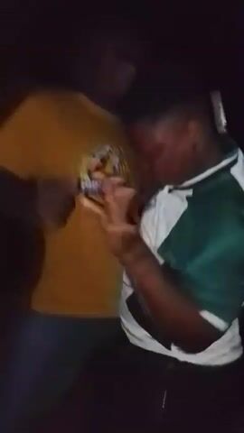 Two males are seen dancing in a dimly lit environment, likely at night in Durumi, Nigeria. One male is wearing a yellow short-sleeved t-shirt with a graphic logo on the front that appears to say "FIRE" with another word below it, possibly "WOLF" or similar. The other male, more prominent in the foreground, is wearing a white and green collared t-shirt, possibly a sports jersey. Both are moving their bodies rhythmically, indicating they are dancing and enjoying themselves. The scene is dark, with only the subjects partially illuminated, contributing to a vibrant, nocturnal atmosphere. The video is slightly blurry due to the low light conditions and movement.