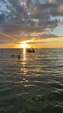 The video shows a serene ocean scene at sunset. The sun is low in the sky, casting a warm golden glow on the water and the clouds. The sky is a mix of orange, yellow, and gray hues, with some clouds obscuring parts of the sun. In the foreground, several people are visible in the water, some with their heads above the surface. In the mid-ground, a small boat, possibly a kayak or canoe, carries a group of people who appear to be paddling. The water is calm with gentle ripples, and the sunlight creates a shimmering reflection on the surface. The overall atmosphere is peaceful and idyllic, characteristic of a sunset at a beach or coastal area.