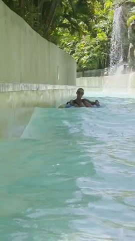 A person is riding an inner tube down a water ride. The person is wearing sunglasses and a dark swimsuit. The water ride has a waterfall in the background. The water ride is surrounded by lush greenery.