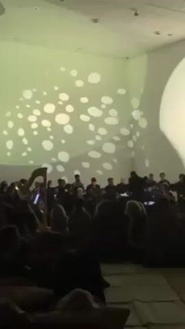 The video shows a dimly lit indoor space, likely a hall or auditorium, where an orchestra is performing. The musicians, dressed in dark clothing, are seated in front of the audience. A conductor, silhouetted, stands facing them, directing the performance. A harp is visible on the left side of the frame. The audience is seated on the floor, many appearing to be leaning against pillows for comfort. The walls are painted white, with a pattern of large, round shapes projected onto them, casting a soft, ambient light. The overall atmosphere is one of relaxed appreciation of the musical performance.