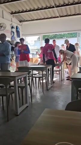 The video shows the interior of a casual dining establishment, possibly a cafe or restaurant. Sunlight streams in through large windows at the back, illuminating the space. Several tables and chairs are visible, some occupied by patrons and others empty. A group of people, likely customers and staff, are gathered around a central table. A young person in a red and blue shirt and white shorts stands on the left, holding a phone. A person in a pink t-shirt with a logo is interacting with customers at the central table, possibly taking orders or serving food. Other individuals, appearing to be tourists, are also present, some looking at menus or engaging in conversation. On the left side of the establishment, racks of merchandise, possibly clothing or accessories, are displayed. A ceiling fan is mounted on the wall. The overall atmosphere appears relaxed and busy.