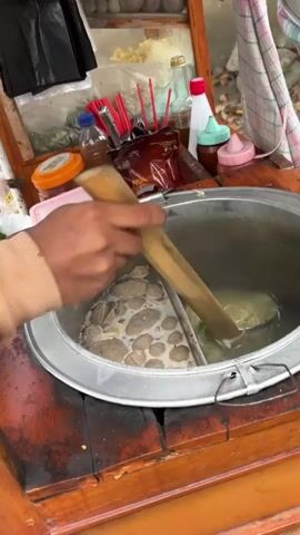 A person's hands are visible, using chopsticks to lift a metal strainer filled with cooked noodles from a large metal pot. The pot is divided into two sections. The left section contains numerous round, brownish meatballs submerged in broth. The right section holds the boiling noodles. The pot is situated on a wooden cart, which appears to be a food stall. Various condiments and utensils are arranged on a shelf behind the pot, including bottles of sauce, red straws, and other food items. The background is slightly blurred but suggests an outdoor setting. The overall scene depicts the preparation of a noodle dish, likely at a street food vendor.