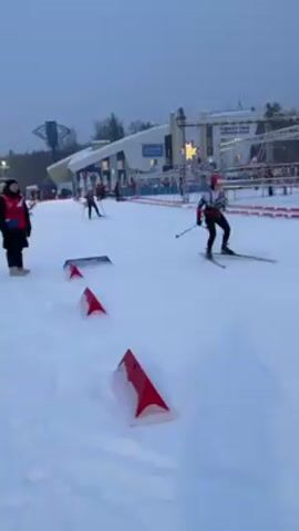 The video shows a cross-country skiing competition or practice session in a snowy outdoor environment. Two skiers are visible, moving from right to left in the frame. The skiers are wearing appropriate skiing gear and using poles for propulsion. Red triangular markers are lined up along the course.  In the background, there is a building with signage in Cyrillic. A large tower structure is visible in the distance. Several people are spectating or involved in the event, as one person in a vest is standing near the camera. The weather appears overcast.
