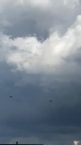 The video shows two helicopters flying in a cloudy sky. The helicopters are small silhouettes against the large, dramatic clouds, one on the left side of the frame and one on the right. The clouds are a mix of dark gray storm clouds and brighter white clouds, suggesting overcast or potentially stormy weather. The bottom of the frame shows a hint of a landscape with a few trees and what appears to be a structure, possibly a building or a lighthouse, in the lower left corner. The helicopters are the primary subjects of focus in the sky.