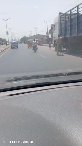 The video shows a street scene in what appears to be a developing country, filmed from the dashboard of a car. The road is paved and divided by white lines.  Several motorized vehicles are visible: a small blue pickup truck, a motorcycle with two people, and several yellow three-wheeled vehicles, also known as auto rickshaws or tuk-tuks. One tuk-tuk is overturned, and several people are standing around it, presumably righting it. There are also some pedestrians visible. The environment includes overhead power lines, streetlights, and buildings along the side of the road. The weather seems bright and sunny. The footage is a bit shaky, as one might expect from a camera mounted in a moving vehicle. A watermark indicating “TECNO SPARK 20” is at the bottom of the video.