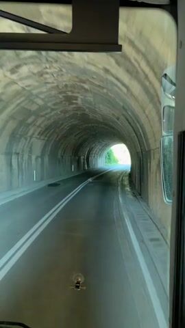 The video shows a point-of-view perspective from inside a vehicle, likely a bus, as it travels through a tunnel and emerges into daylight. The tunnel is constructed of rough-hewn stone or concrete, with a distinctly arched ceiling. The road within the tunnel is wet, with tire tracks visible on the asphalt and reflections of the light from the tunnel's exit shimmering on the surface, suggesting recent rain. Double white lines mark the lanes of the road.

As the vehicle progresses, the end of the tunnel comes into view, revealing a bright, washed-out daylight scene with green foliage on either side of the road. The transition from the dark, enclosed tunnel to the open, sunlit exterior is stark. The side of the vehicle, including a mirror and window, is visible along the right edge of the frame, indicating the perspective is from the driver's or a front passenger's seat. A small, dark object, possibly a dashcam mount, is centered on the dashboard.

The overall impression is one of movement and transition, moving from the confined space of the tunnel to the open road. The wetness of the road and the bright daylight suggest it might be a sunny day following a period of rain. The presence of lush greenery hints at a natural, possibly mountainous, environment. No people are explicitly visible, but the activity of driving through a tunnel and emerging into daylight is the primary event. There is no visible text in the video. The implied location of Prijevorac, Montenegro, would further support the idea of a scenic, potentially mountainous route.