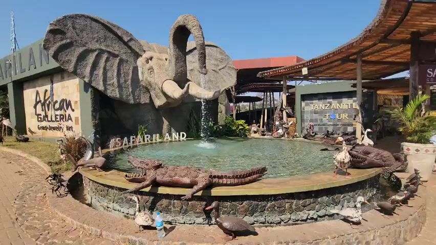 The scene depicts a fountain and surrounding decorative structures at the entrance of "Safariland" in Tanzania, Africa. A large, sculpted elephant head serves as the main feature of the fountain, with water flowing from its trunk into a circular pool. A sculpture of a crocodile lies on the edge of the pool, and various waterfowl statues, mostly geese and ducks, are scattered around the fountain. The background features two building structures, "African Galleria" and "Tanzanite Tour" which appear to be shops or businesses. The setting is outdoors, with a bright, clear sky. The ground surrounding the fountain is paved.