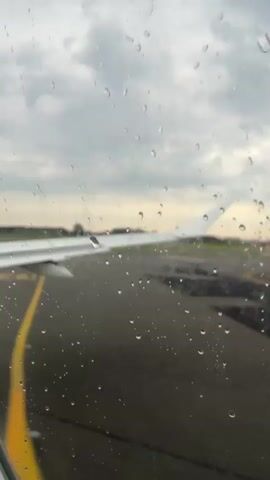 A video taken from inside an airplane looking out the window. Raindrops are visible on the window. The wing of the airplane is visible on the left side of the frame. The runway is visible in the distance, with yellow markings. The sky is overcast and gray.
