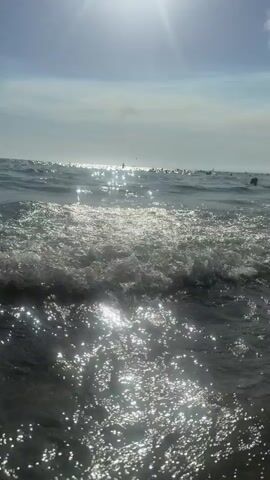 The video shows a close-up view of ocean waves under a bright sky with the sun reflecting off the water. The camera appears to be at water level, capturing the movement of the waves and the shimmering sunlight. In the distance, several dark shapes that could be people or buoys are visible in the water. The sky is partly cloudy with the sun positioned high and to the left of the frame. The overall scene depicts a sunny day at the beach or a similar coastal environment.