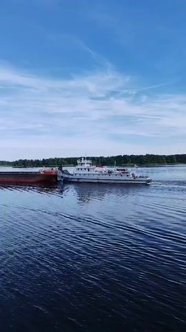 A white boat pushes a long red barge on a wide body of water, from right to left, under a blue sky with scattered clouds. Trees line the distant shore.