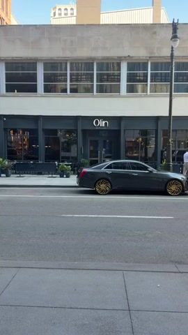 The video pans across the side of a dark gray luxury sedan parked on a city street. The car has distinctive, large, gold-colored custom wheels with a swirling spoke design. Behind the car is the facade of a building with large windows and a dark gray entrance. Above the entrance, a white illuminated sign reads "Olin Bar + Kitchen". There are outdoor seating arrangements visible to the left of the entrance, including tables and chairs. The street in front of the car appears to be paved, with a sidewalk to the left. The building has multiple floors, with the upper floors featuring large, multi-paned windows. A streetlight is visible to the right of the building. The lighting suggests it is daytime, possibly late afternoon or early evening.