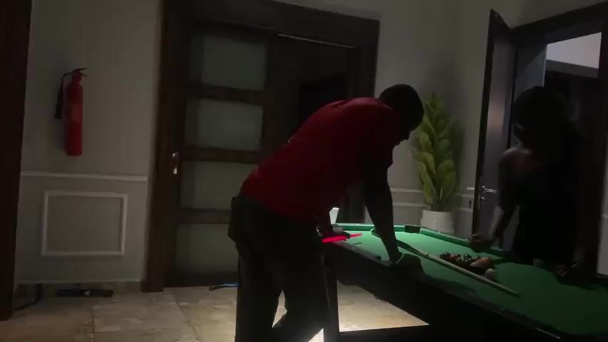 The video shows two people, a man and a woman, standing on opposite sides of a pool table in a room. The man, wearing a red shirt and dark pants, is on the left side of the table, bending over as if to take a shot. He has a pool cue in his hands. The woman, wearing a black dress, is on the right side of the table, looking at the balls. The pool table is green with a set of colored balls arranged in a triangle. There are two pool cues on the table. In the background, there is a large doorway with glass panels and a red fire extinguisher mounted on the wall to the left. A large potted plant is visible behind the pool table. The room appears to be dimly lit, with a single light source from above casting shadows. The walls are decorated with white molding.