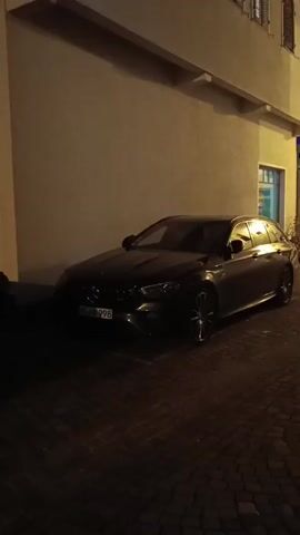 A dark colored Mercedes-Benz wagon is parked on a cobblestone street at night, next to a light brown building. The car's headlights are on and illuminate the license plate which reads "WN NB 998". The car is parked in a marked parking space. The building appears to have a window visible on the right side of the frame.