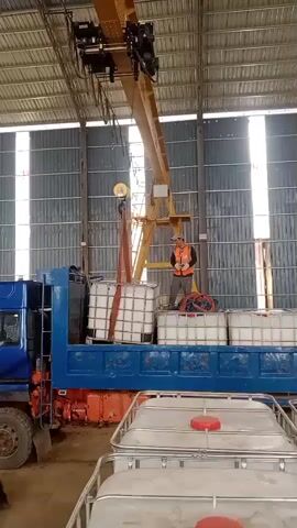 A man in a high-visibility vest and hard hat operates a remote control for a yellow overhead crane. The crane is lifting a large white plastic tote bin, suspended by orange straps, from the bed of a blue flatbed truck. The truck is positioned inside a large warehouse-like building with corrugated metal walls and a metal ceiling. The truck bed is loaded with several more white tote bins. In the foreground, two more white tote bins are stacked. The man is standing on the truck bed, overseeing the operation.