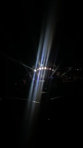 The video shows a dark stage with bright spotlights shining from above. The lights create a lens flare effect. There are several people visible in the background, appearing to be an audience. A large circular screen is positioned on the stage behind the lights. The overall atmosphere suggests an indoor event, possibly a concert or performance.
