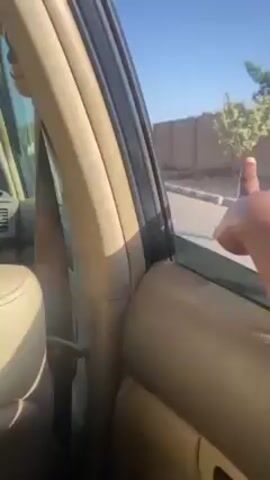 The video shows an arm extending from a car window, the hand held in an open position, as the car drives along a street lined with walls and buildings under a bright, blue sky. The color of the interior of the car is beige. Electrical poles and lines are visible along the roadside.