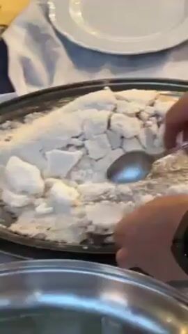 Here is a description of the video:
A person is using two spoons to break apart a large, salt-covered fish on a silver platter. The fish is completely encased in the salt, which appears to be forming a hard crust. The person is working to crack the salt crust and remove it from the fish. There is a white tablecloth underneath the platter. A white plate and a stainless steel bowl are also on the table.