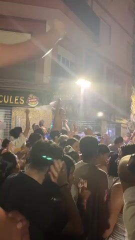 A nighttime outdoor street party is in progress. A crowd of people, densely packed, are visible from the chest up. Many are holding up their phones, suggesting they are recording or photographing. Some individuals are dancing and appear to be celebrating, with hands raised. Water is being sprayed into the air and onto the crowd, creating a misty atmosphere. In the background, buildings with windows and signs are illuminated. One sign reads "JULES". Another sign is partially visible with illuminated letters. The lighting is a mix of streetlights and neon, creating a vibrant, energetic, and somewhat chaotic scene.