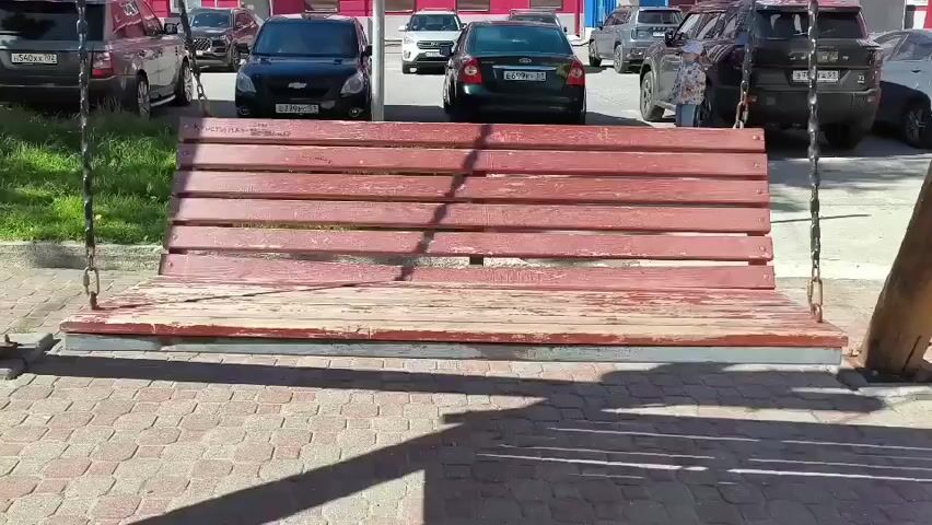 A wooden bench swing hangs from chains, casting a shadow on a paved area. Cars are parked in the background, and a person is visible standing behind the swing, partially obscured. The swing is red and weathered. The environment appears to be outdoors on a sunny day.