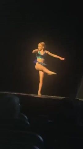 A young girl with blonde hair in a ponytail, wearing a blue and purple leotard, performs a dance routine on a stage. She is lit by a spotlight against a dark background. In the foreground, the dark silhouettes of audience members are visible. The girl executes a leap, followed by a backflip, and then strikes a pose.