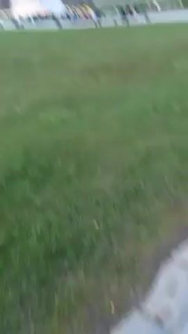The video shows a large outdoor art installation featuring two giant horse heads made of metal. People are walking around the area, some are near the sculptures, and others are walking towards a nearby cafe building. The ground is covered in green grass and patterned pavement. The sky is overcast.