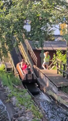 A log flume ride in an amusement park is shown from a high angle.  A log boat carrying five people is moving down a steep, wet track. The people in the boat are looking forward or down. A person in a yellow vest and white shorts is standing on a platform to the right of the track, facing away from the boat and touching their hair. Trees with green leaves surround the ride, and a body of water is visible at the bottom of the track.
